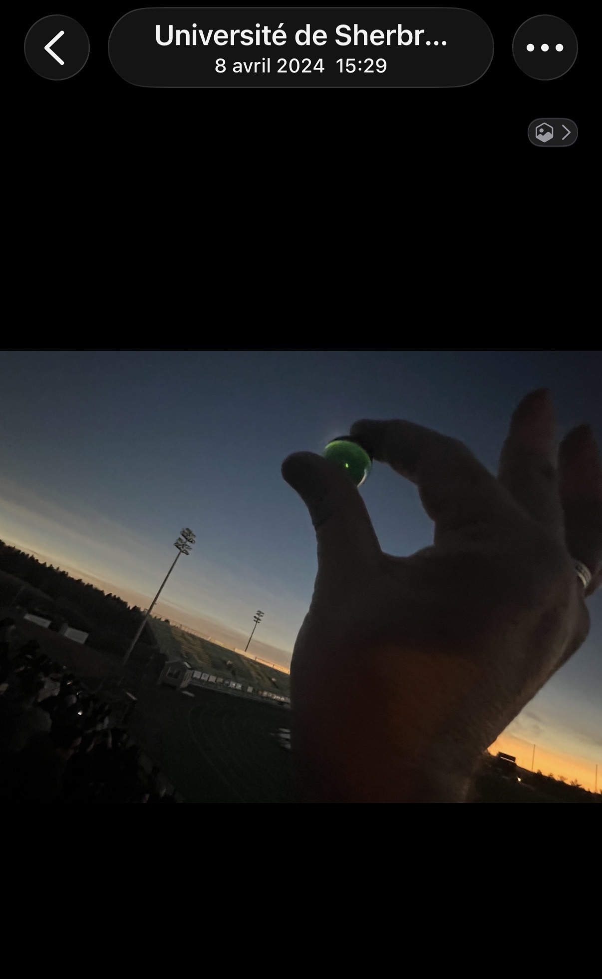 La Perle Verte tenue à bout de doigts contre le ciel crépusculaire de l'éclipse, luisant d'un vert propre au-dessus du stade assombri.