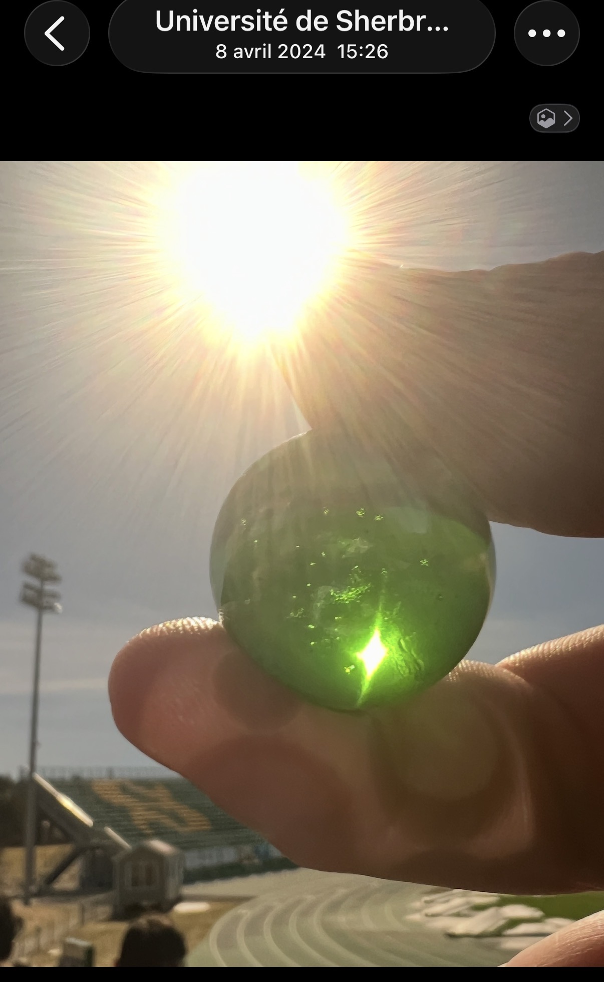 La Perle Verte translucide tenue entre le pouce et l'index, traversée par la lumière éclatante du soleil au-dessus du stade de l'Université de Sherbrooke.